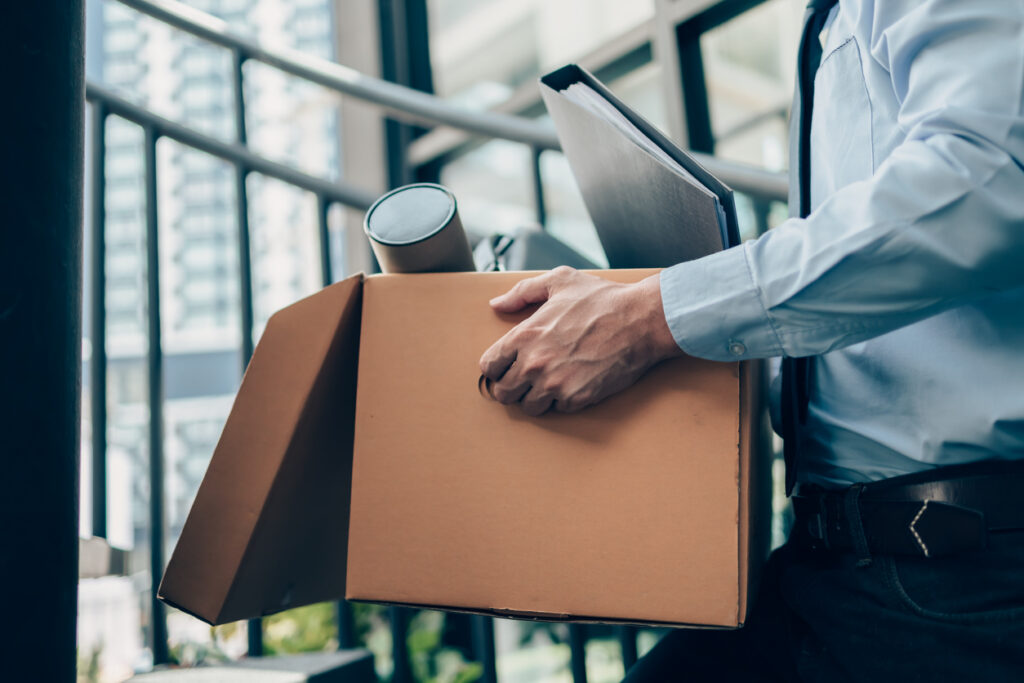 Businessman leaving job carrying box of office belongings including a binder and a tube.